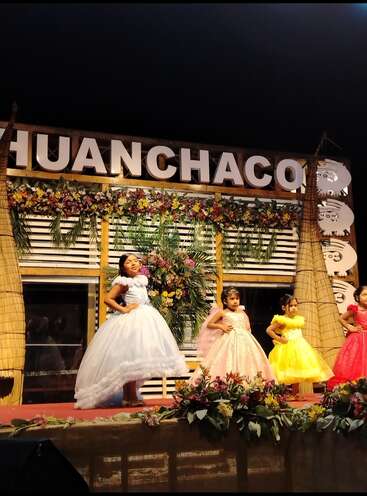 Five young girls in colorful, elegant dresses pose on a decorated stage in Huanchaco, surrounded by flowers and traditional caballitos de totora under night sky.