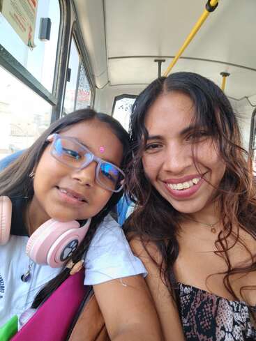 A woman and a young girl smile for a selfie on a bus. The girl wears glasses and pink headphones; both appear happy and close together.