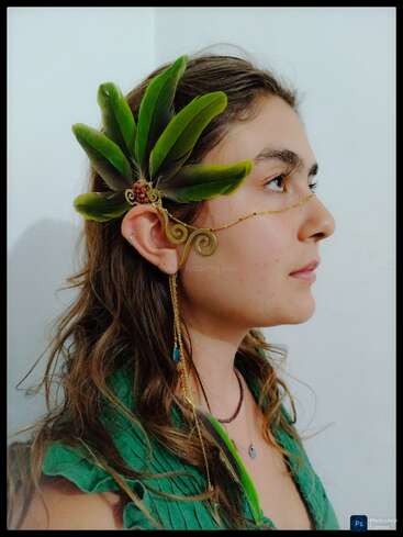 A woman with wavy brown hair wears an elaborate green feather headdress and golden wire accessory that wraps around her ear and extends delicately across her face.