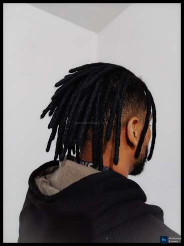 A man with short sides and long black dreadlocks wears a black hoodie. He stands facing a plain white wall, with his back partially turned.