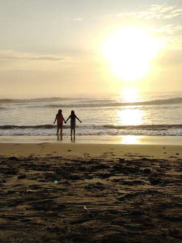 Two people stand hand in hand at the water’s edge, silhouetted by the golden sunset, as gentle waves roll onto the sandy, rocky beach.