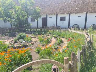Un jardin rustique avec des fleurs colorées et des chemins en briques est entouré d'une clôture en bois. Un vieux bâtiment, un arbre et des roues de chariot se trouvent à l'arrière-plan.
