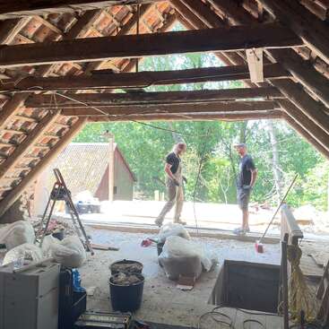 Deux personnes travaillent à un projet de rénovation dans un grenier dont les poutres et les tuiles sont apparentes. Des outils de construction, des sacs et des seaux sont éparpillés sur le sol poussiéreux.