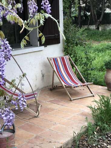Zwei gestreifte Liegestühle stehen auf einer gefliesten Terrasse neben einem weißen Haus. In der Nähe hängen violette Glyzinienblüten. Ein großer Terrakottatopf und grünes Gras umgeben den Bereich.