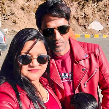 Ein Paar macht an einem sonnigen Tag ein Selfie im Freien. Beide tragen passende rote Jacken und Sonnenbrillen. Vorne ist teilweise ein Kind zu sehen.