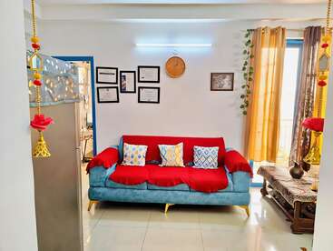 A cozy living room with a blue sofa, colorful cushions, wall art, certificates, hanging decorations, potted plant, and sunlight streaming through golden curtains. Neat and inviting.