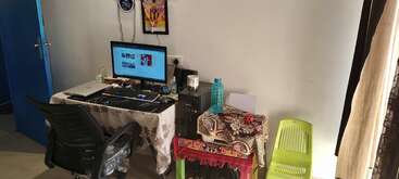 A home workspace with a computer, keyboard, mouse, and monitor on a table covered with a cloth. There’s a chair, green stool, water bottle, and decor.