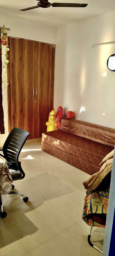 A sunlit room with white walls, brown wardrobe, brown sofa, office chair, decorative soft toys, ceiling fan, and patterned chair, creating a cozy environment.