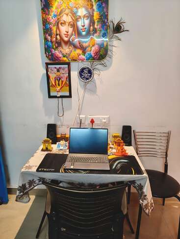 Un espace de travail avec un ordinateur portable posé sur une table, des images religieuses, des objets décoratifs, une plume de paon, une nappe à motifs floraux, des haut-parleurs, une chaise et une atmosphère spirituelle.