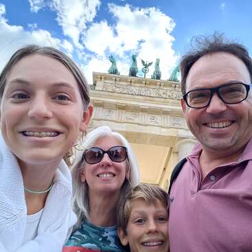 The image shows a family of four posing for a selfie in front of the Brandenburg Gate in Berlin, Germany, with a blue sky and white clouds in the background.