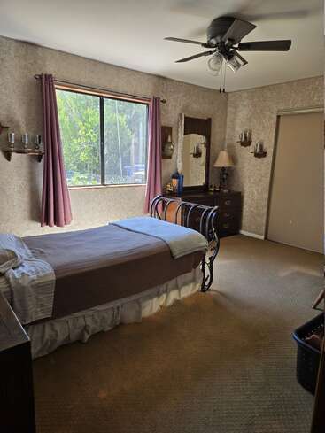 Ein gemütliches Schlafzimmer mit einem Einzelbett, brauner Tagesdecke, großem Fenster mit lila Vorhängen, Deckenventilator, Kommode mit Spiegel, Lampe und Wandregalen.