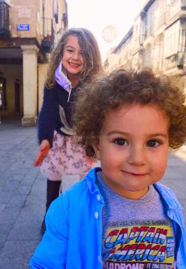 Das Bild zeigt zwei Kinder, die auf einer Straße in der Stadt stehen. Der Junge im Vordergrund trägt ein blaues Hemd und das Mädchen hinter ihm eine lila Jacke.