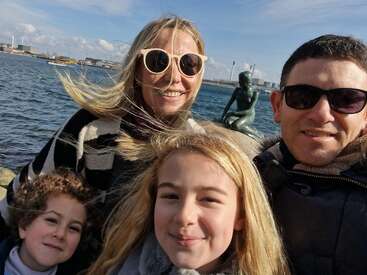 Das Bild zeigt eine vierköpfige Familie, die für ein Selfie vor einem Gewässer posiert, mit der Statue einer Meerjungfrau im Hintergrund.
