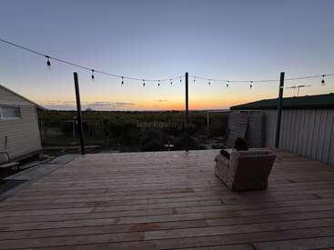 Une personne est assise seule dans un fauteuil sur une terrasse en bois au coucher du soleil, entourée de guirlandes lumineuses, face à un magnifique paysage et à un ciel dégagé. L'atmosphère est paisible et sereine.