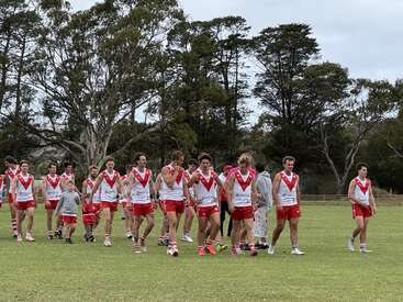 Un groupe de joueurs de football australien, vêtus d'uniformes rouges et blancs, traverse un terrain en herbe, entouré d'arbres sous un ciel nuageux, l'air détendu.