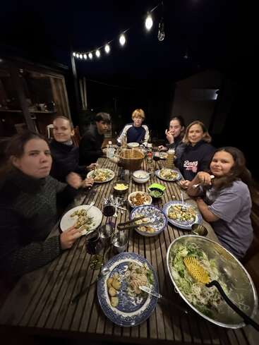 Siete personas están reunidas alrededor de una mesa de madera al aire libre por la noche, cenando juntas bajo unas guirnaldas de luces. Se ven platos de comida y ensaladeras.
