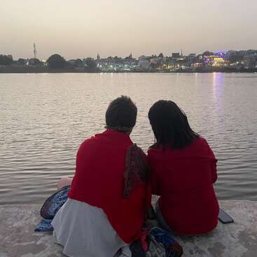 Deux personnes sont assises sur une corniche en pierre au bord d'une grande étendue d'eau, avec un horizon urbain en arrière-plan, sous un ciel brumeux.