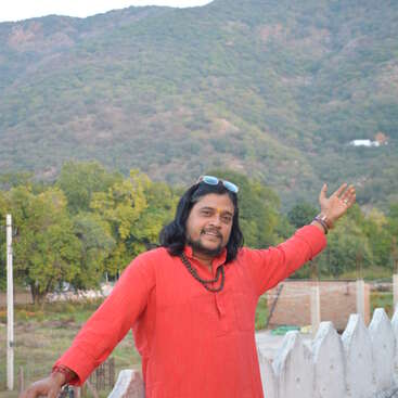 A man in a red kurta, wearing beads and sunglasses on his head, smiles and gestures proudly, standing outdoors with lush green hills in the background.