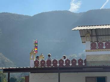 Trois personnes se tiennent sur un toit orné d'une balustrade rouge et de décorations dorées. Des drapeaux colorés flottent à leurs côtés, avec des montagnes brumeuses formant une toile de fond sereine.