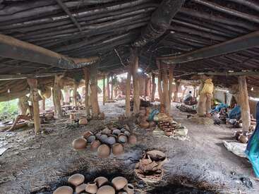 Esta imagem mostra uma oficina de cerâmica tradicional com um telhado de palha. Potes e vasos de barro estão espalhados, enquanto várias pessoas trabalham em várias tarefas de cerâmica.