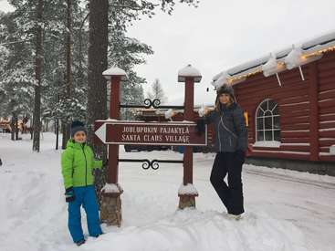 The image depicts a woman and a child standing beside a signpost in front of a log cabin, with a snowy landscape and trees in the background.