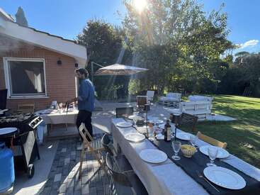 An einem sonnigen Tag grillt ein Mann vor einem Haus. Ein langer Esstisch ist mit Tellern und Gläsern gedeckt und bereit für ein Essen im Freien.