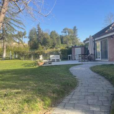 Ein Steinweg schlängelt sich durch einen grünen Rasen hin zu einer Terrasse mit Sitzgelegenheiten, Sonnenschirm und Tisch, umgeben von Bäumen und einem modernen Haus an einem sonnigen Tag.