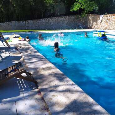 Kinder spielen und planschen in einem strahlend blauen Swimmingpool. Sonnenlicht und Schatten fallen auf den Poolbereich, wo Stühle und Spielsachen verstreut liegen und eine fröhliche Szene ergeben.