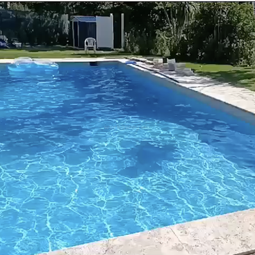 Ein glitzernder blauer Pool liegt im strahlenden Sonnenlicht, umgeben von Grün und Steinfliesen. Im Hintergrund stehen ein leerer weißer Stuhl und eine blaue Schwimmhilfe.