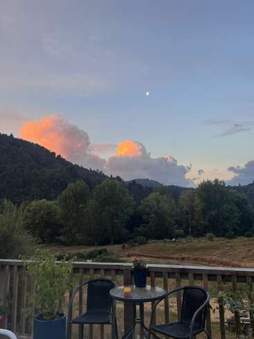 A peaceful outdoor patio with two chairs and a table overlooks a green landscape, trees, and hills. Orange clouds and a crescent moon decorate the evening sky.