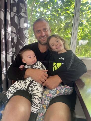 A smiling man sits indoors with two young children on his lap, surrounded by leafy green plants and natural light from a large window behind them.