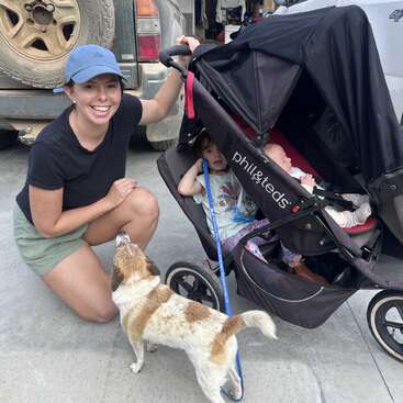 A smiling woman kneels beside a double stroller with two children inside. A small, brown and white dog on a leash looks up at her.