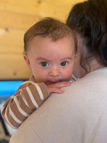 A baby with big, curious eyes rests on an adult’s shoulder, wearing a brown and white striped outfit. The background is softly lit and wooden.