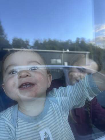 A smiling baby presses its face and hand against a window, with an older child behind. Blue sky and trees are reflected outside in the glass.