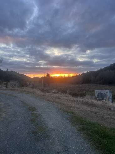 A gravel path leads through grassy fields at dusk. The sun sets behind distant hills, painting the clouds with warm orange and purple hues, creating a serene landscape.