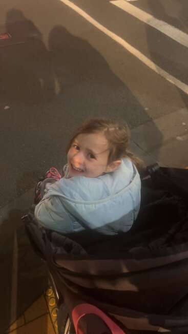 A young girl in a light blue coat sits smiling in a stroller at night on a sidewalk, with street lights casting warm shadows nearby.