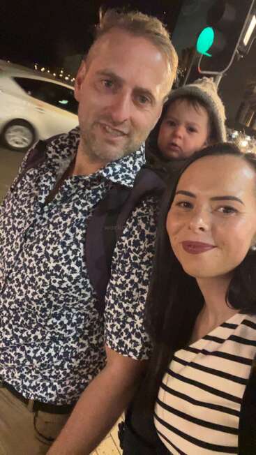 A man, woman, and child are smiling at night outdoors. The child is in a carrier on the man’s back. A car passes behind them.