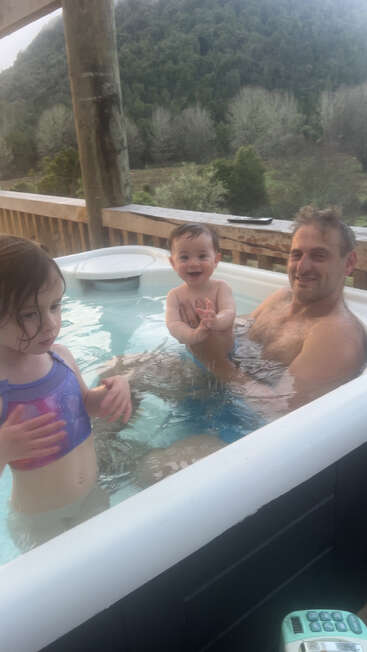 A man and two young children enjoy relaxing in a hot tub outdoors, surrounded by natural greenery and hills on a peaceful, wooden deck.