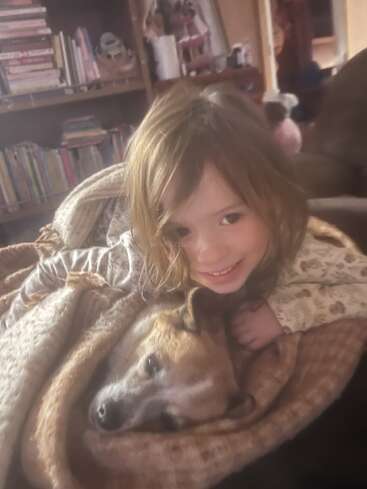 A smiling child cuddles closely with a relaxed dog, both wrapped in a cozy blanket. Bookshelves are visible in the warm, softly-lit background room.