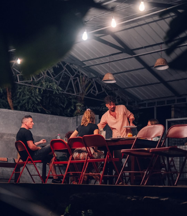 Four people sit and converse around a table under a metal-roofed structure. Red chairs, warm lighting, trees, and a relaxed, evening outdoor dining atmosphere are present.