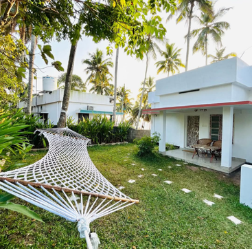 A peaceful garden with a hammock, surrounded by lush greenery and palm trees. White house with a porch and outdoor seating create a relaxing atmosphere.