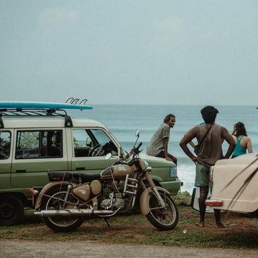 Four people are relaxing near the ocean, gathered by a motorcycle and a van with surfboards on top, enjoying a casual, beachside atmosphere together.