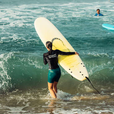 A woman carrying a yellow surfboard enters the ocean, wearing a black rash guard and green shorts. Another person is seen swimming in the background. Waves crash.