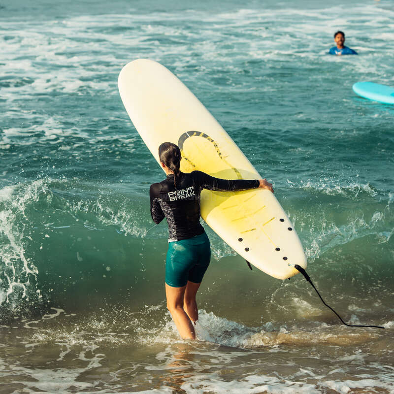 A woman carrying a yellow surfboard enters the ocean, wearing a black rash guard and green shorts. Another person is seen swimming in the background. Waves crash.