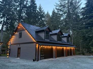 Eine moderne zweistöckige Garage mit drei Toren, Dachgauben und Lichterketten außen, umgeben von hohen Kiefern und an einer Kiesauffahrt in der Abenddämmerung.