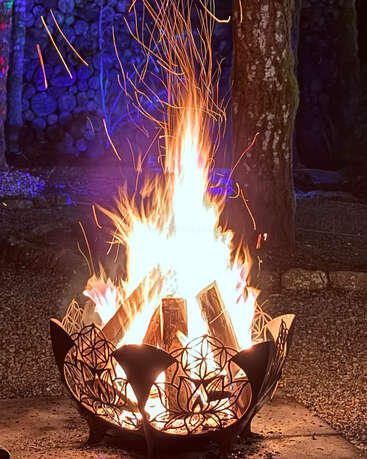 Eine dekorative Feuerstelle mit aufwendigen Ausschnittmustern lodert hell und lässt Funken in die Höhe steigen. Umgeben von Kies, Bäumen und Holzscheiten sorgt das leuchtende Feuer für eine gemütliche Atmosphäre.
