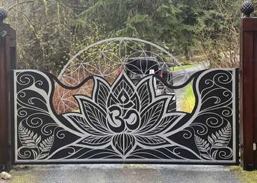 Ein dekoratives Metalltor mit einer Lotusblüte, dem Om-Symbol, wirbelnden Mustern, Blättern und geometrischen Formen, das vor dem Eingang einer von Bäumen gesäumten Auffahrt steht.