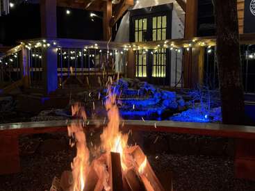 Eine gemütliche Szene im Freien bei Nacht mit einem lodernden Lagerfeuer, Holzbänken, Lichterketten und einem warm beleuchteten Haus, dessen Veranda mit festlicher Beleuchtung geschmückt ist.