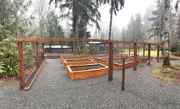 Dieses Bild zeigt einen umzäunten Garten mit mehreren erhöhten Holzbeeten, umgeben von Bäumen, Kieswegen und einem nebligen Wald im Hintergrund an einem bewölkten Tag.