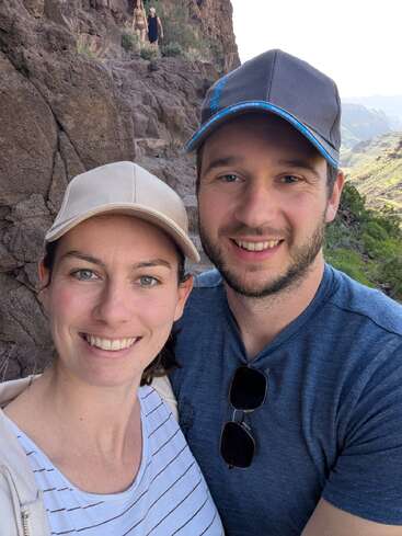Una pareja sonriente con gorras se hace un selfi durante una excursión por un sendero rocoso de montaña. Al fondo se ve a otra pareja de excursionistas disfrutando de la naturaleza.